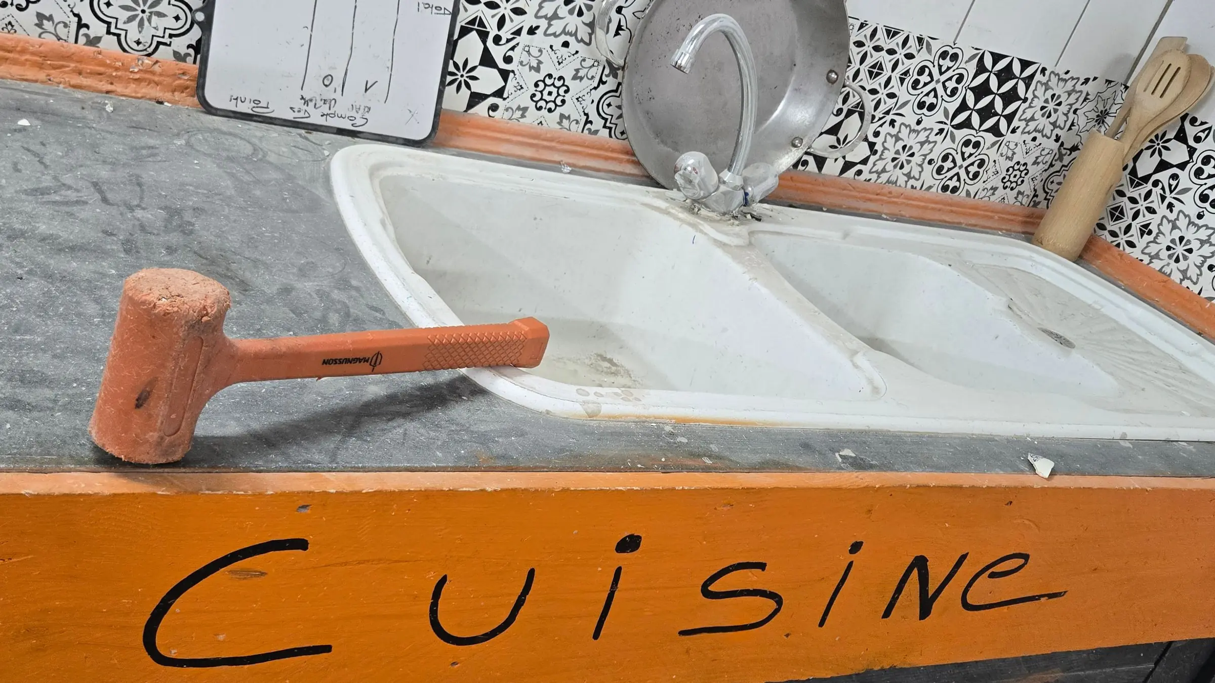 Un maillet en caoutchouc posé sur le plan de travail de la salle cuisine, devant un vieil évier et le mot Cuisine peint à la main sur une planche orange