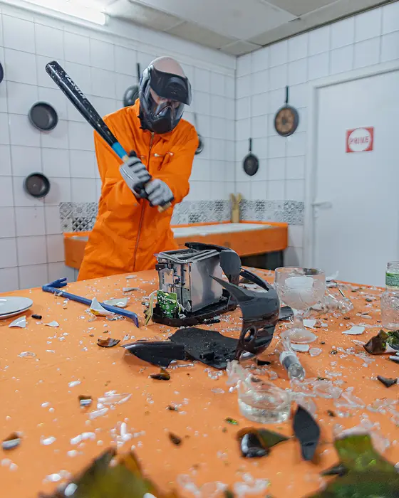 Batte levée sur un grille-pain, la table jonchée de verre brisé dans la salle cuisine de la rage room AtelierKS