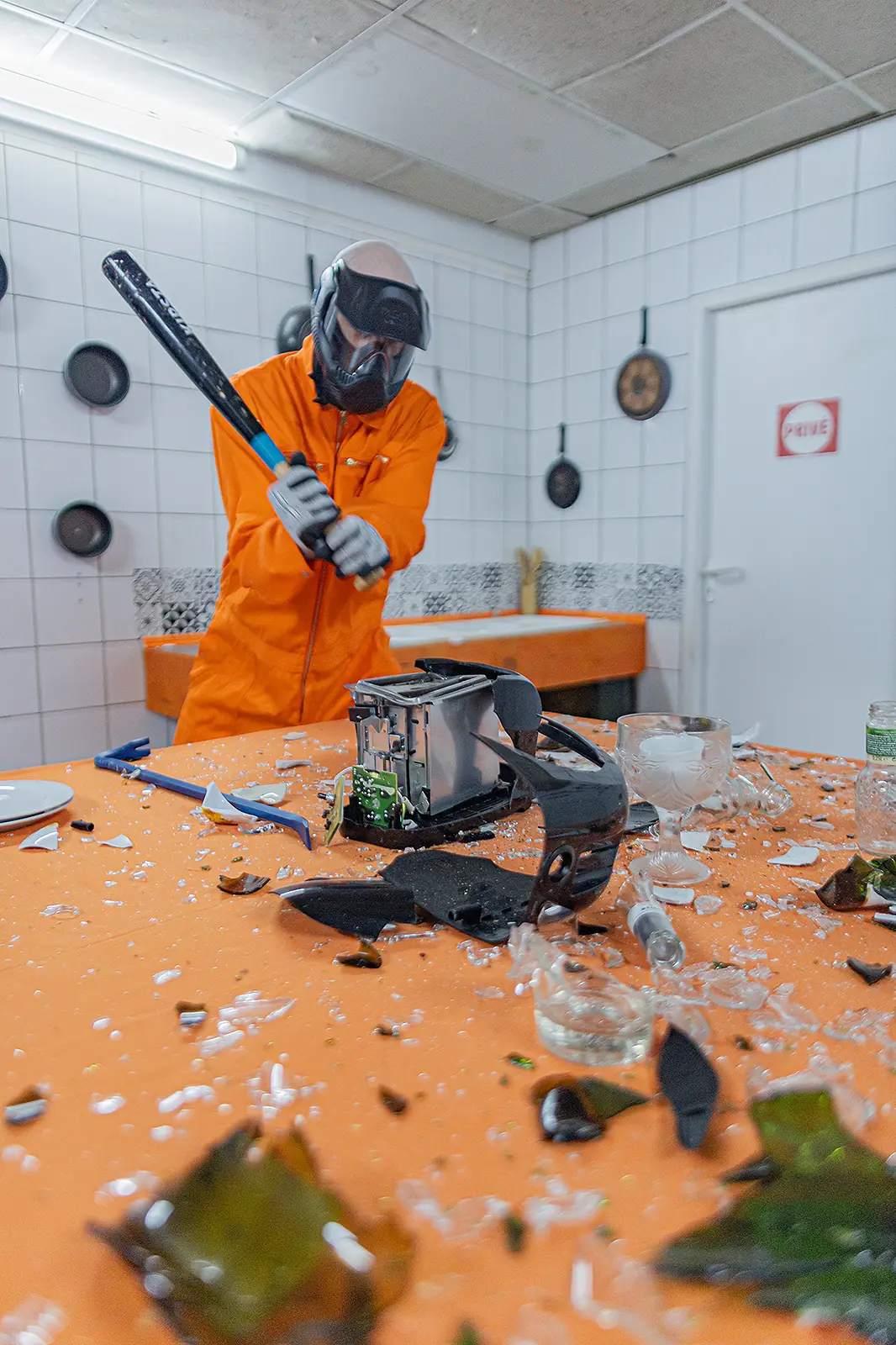 La cuisine de l'AtelierKS pendant une séance : une
personne en combinaison orange et casque à visière lève
une batte de baseball sur un grille-pain, sur une table
jonchée de verre brisé