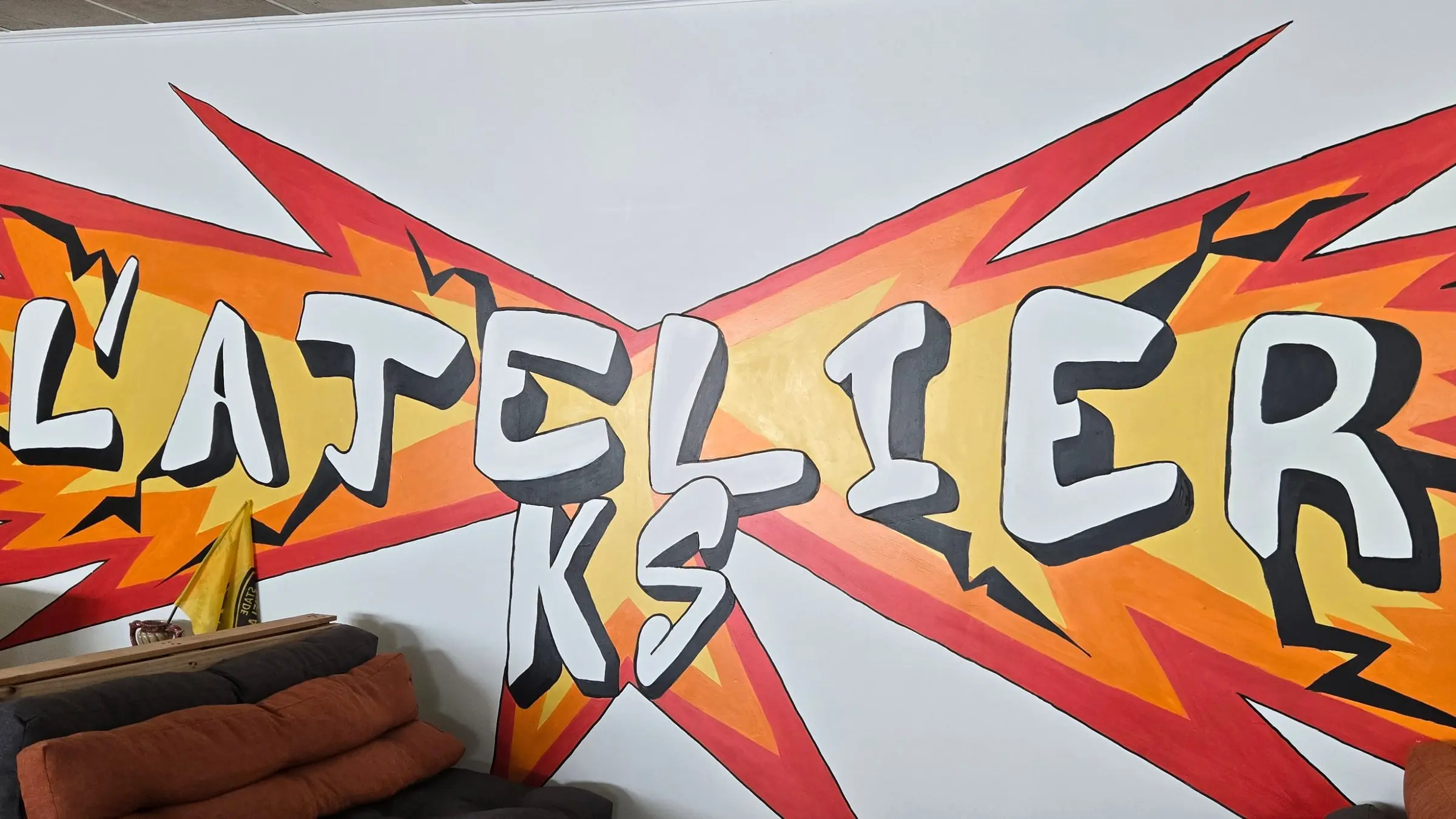 Le nom L'ATELIER KS peint en lettres blanches sur un éclair rouge et orange, sur le mur de la salle principale de l'atelier de destructothérapie de La Rochelle