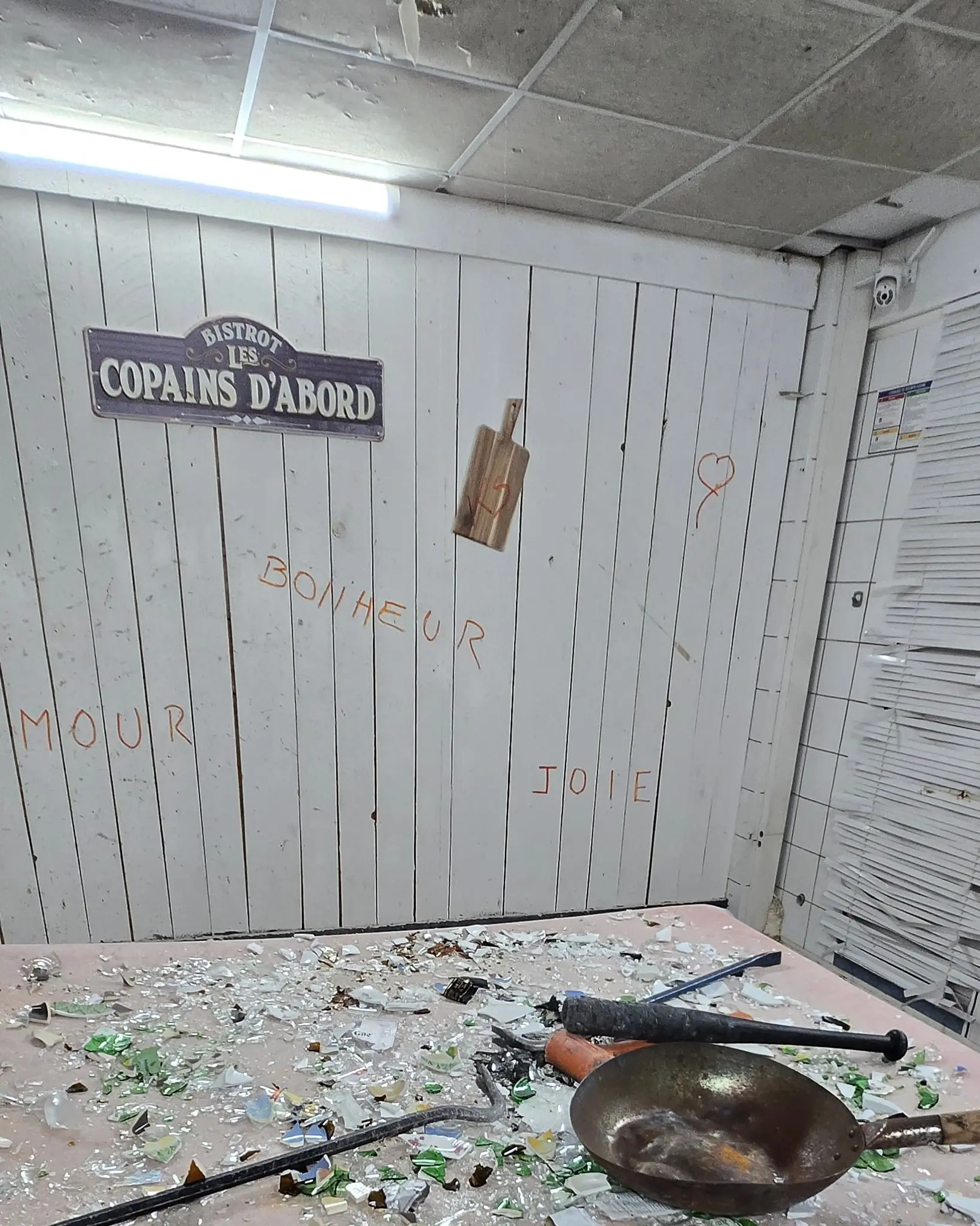La cuisine de l'AtelierKS après une séance : table couverte d'éclats de verre et de porcelaine, poêle en fonte, masse et pied-de-biche, et les mots AMOUR, BONHEUR, JOIE écrits en orange sur le mur en lambris blanc