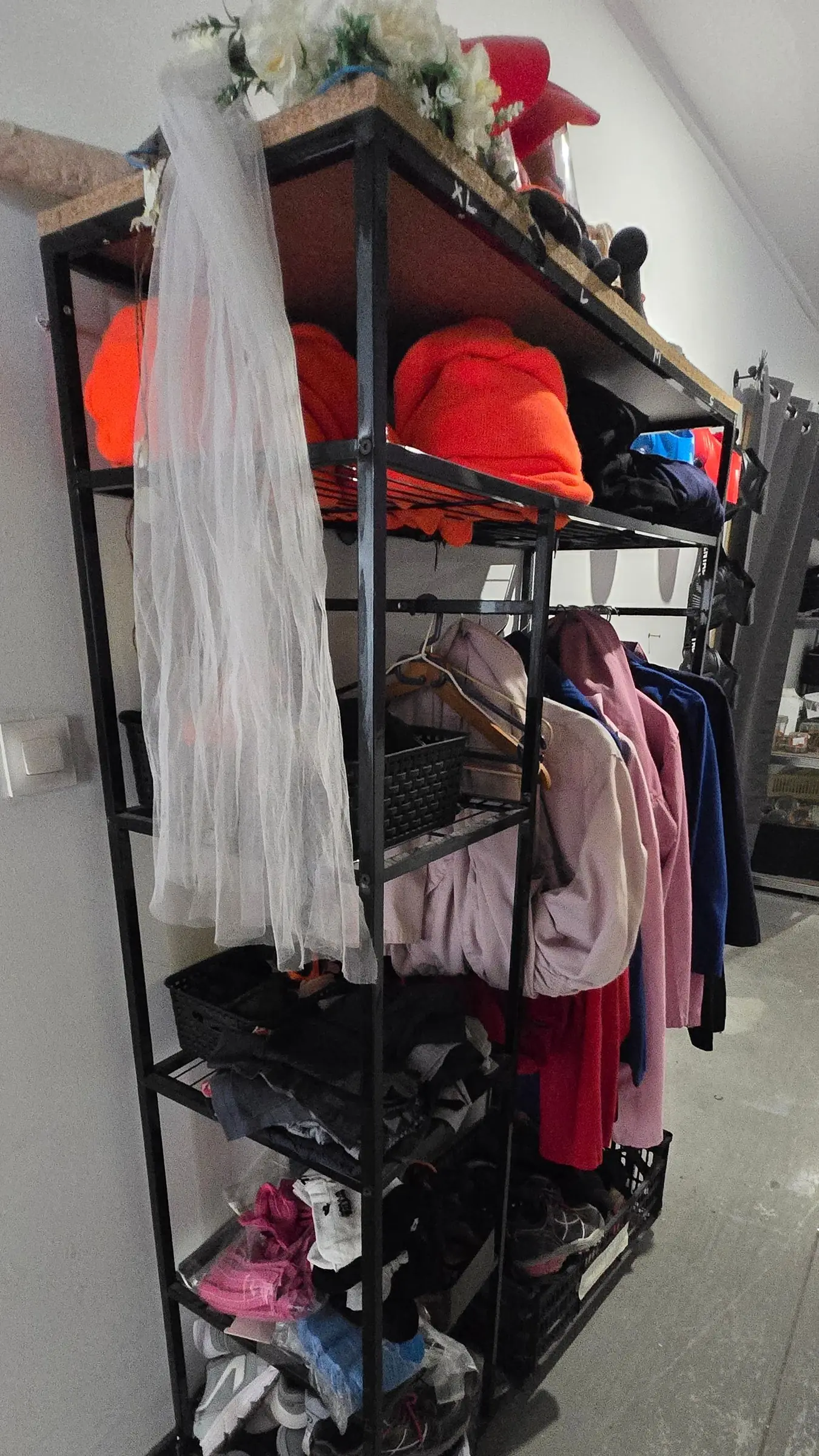 Étagère du vestiaire de la salle de défoulement :
combinaisons rose, bleue et rouge sur cintres,
bonnets orange, chaussures fermées en bacs, et un
voile de mariée accroché sur le côté