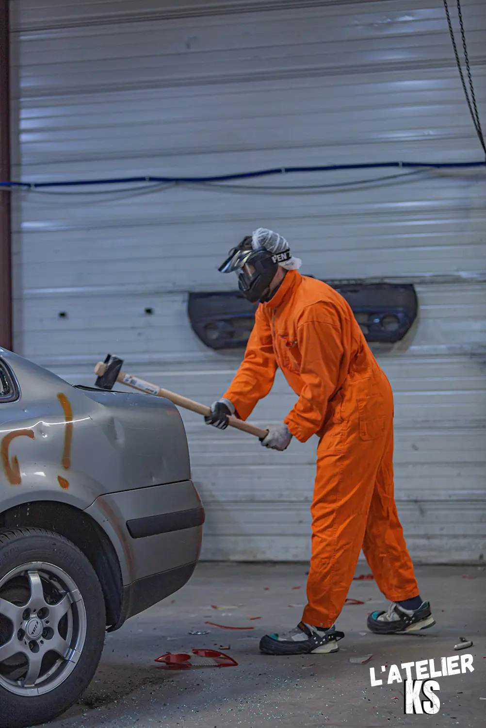 Le garage de l'AtelierKS : une personne en
combinaison orange frappe à la masse l'aile arrière
d'une voiture grise taguée, devant un rideau métallique